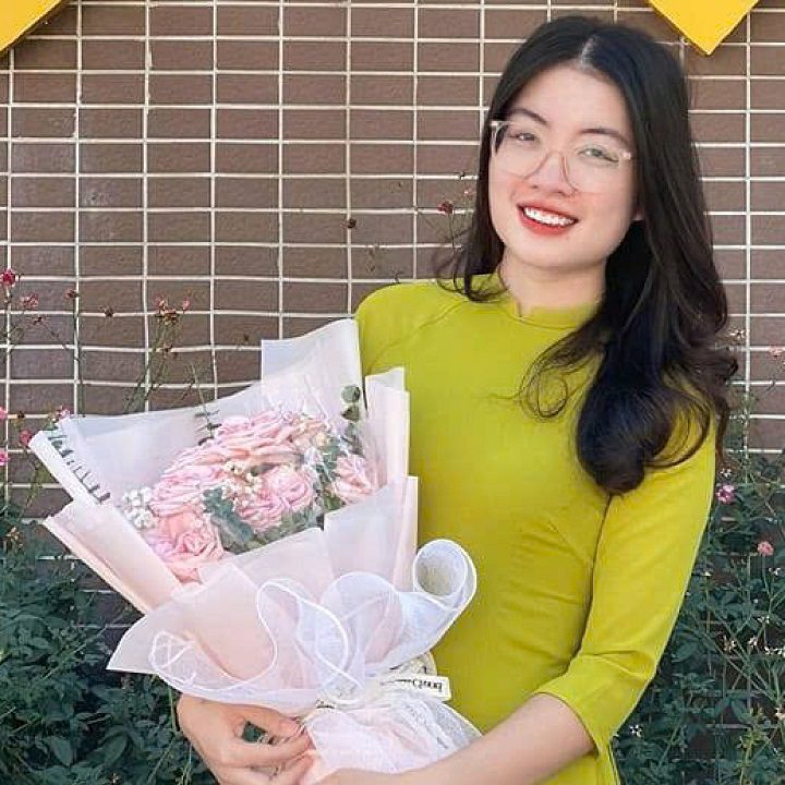 Cô gái trẻ mỉm cười rạng rỡ mặc bộ áo dài truyền thống màu xanh cốm, tay cầm bó hoa hồng phấn lớn, đeo kính trắng, tóc đen dài uốn lượn sóng, chụp ảnh kỷ niệm trên nền tường gạch.